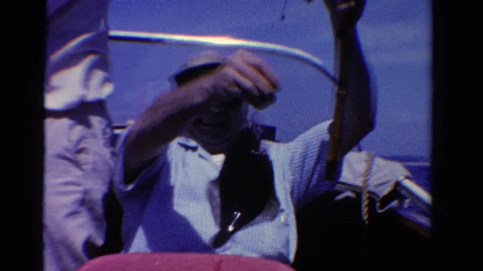 1958 Ontario home-movie frame with a seated adult man in a cap handling a fishing line while another person stands nearby on the boat.