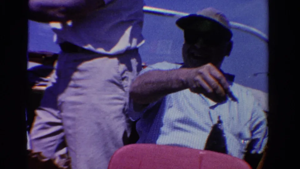 1958 Ontario home-movie frame showing a seated man in a cap presenting a small fish on a line from a boat in bright daylight.