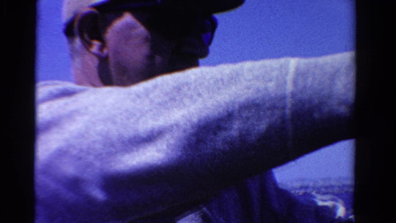 Man in a cap and light sweater stands on a boat with blue water behind him.