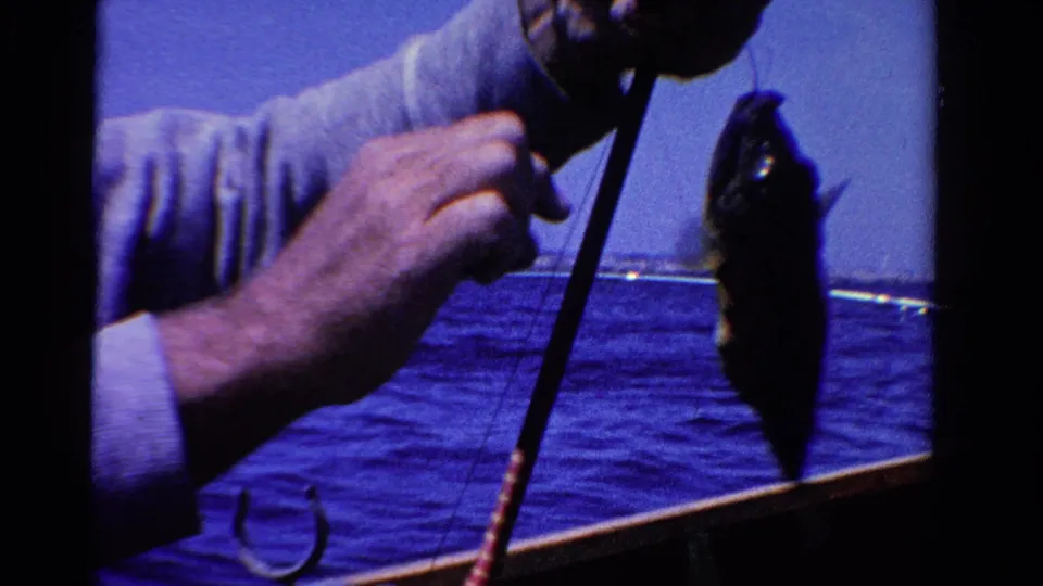 1958 Ontario home-movie frame with a man’s hands and fishing rod as a small fish hangs on the line above blue water.
