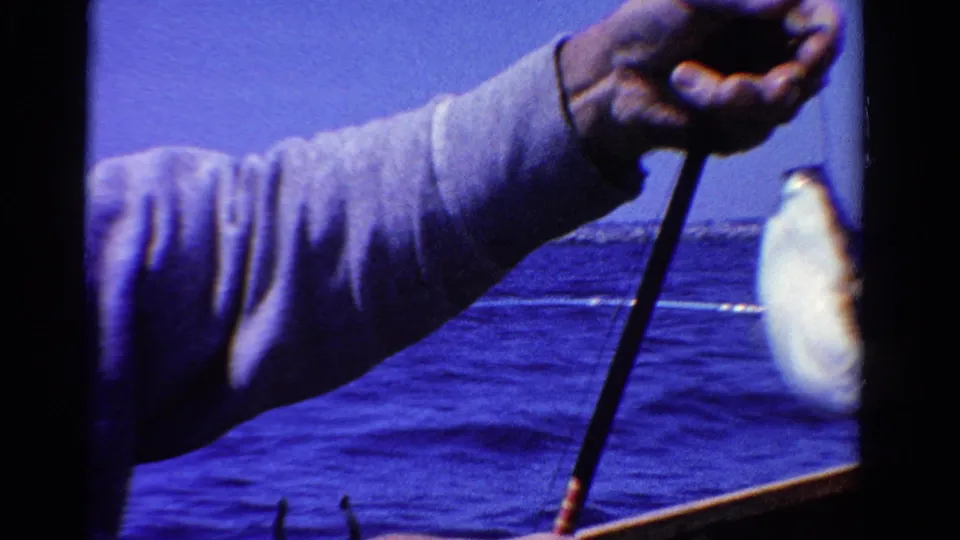1958 Ontario home-movie frame of a small fish suspended from a line beside the man’s arm and fishing rod, with open water behind.