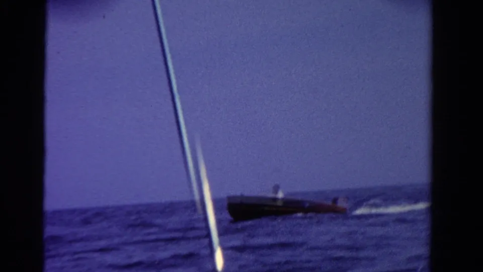 1958 Ontario home-movie frame with a distant man piloting a dark motorboat across a calm lake under a blue sky.