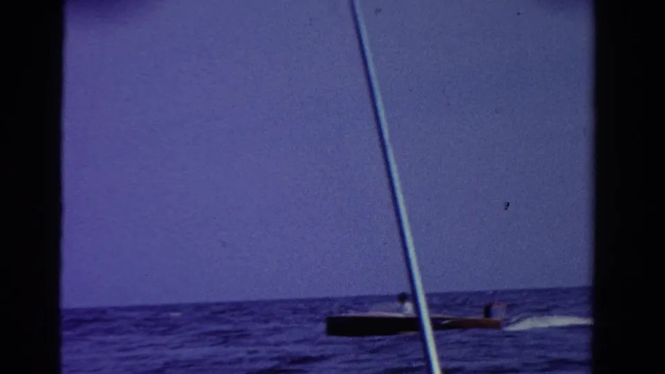 1958 Ontario 8mm frame showing a dark motorboat low on the lake horizon, partly obscured by a diagonal foreground pole.