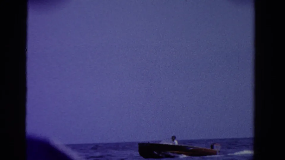 1958 Ontario home-movie frame with a distant man in a dark motorboat speeding along the lower edge of a blue lake scene.