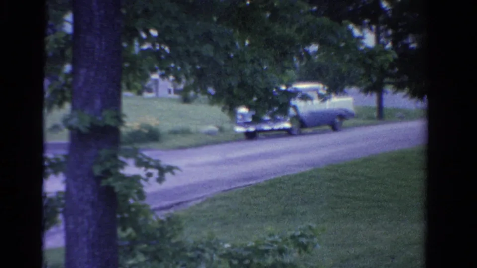 A mid-century car drives along a tree-lined Ontario road, partly framed by foliage and roadside grass.