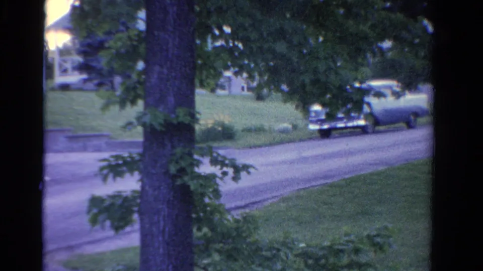 1958 Ontario 8mm view of a quiet residential street with mature trees, lawns, houses, and a car moving along the road.