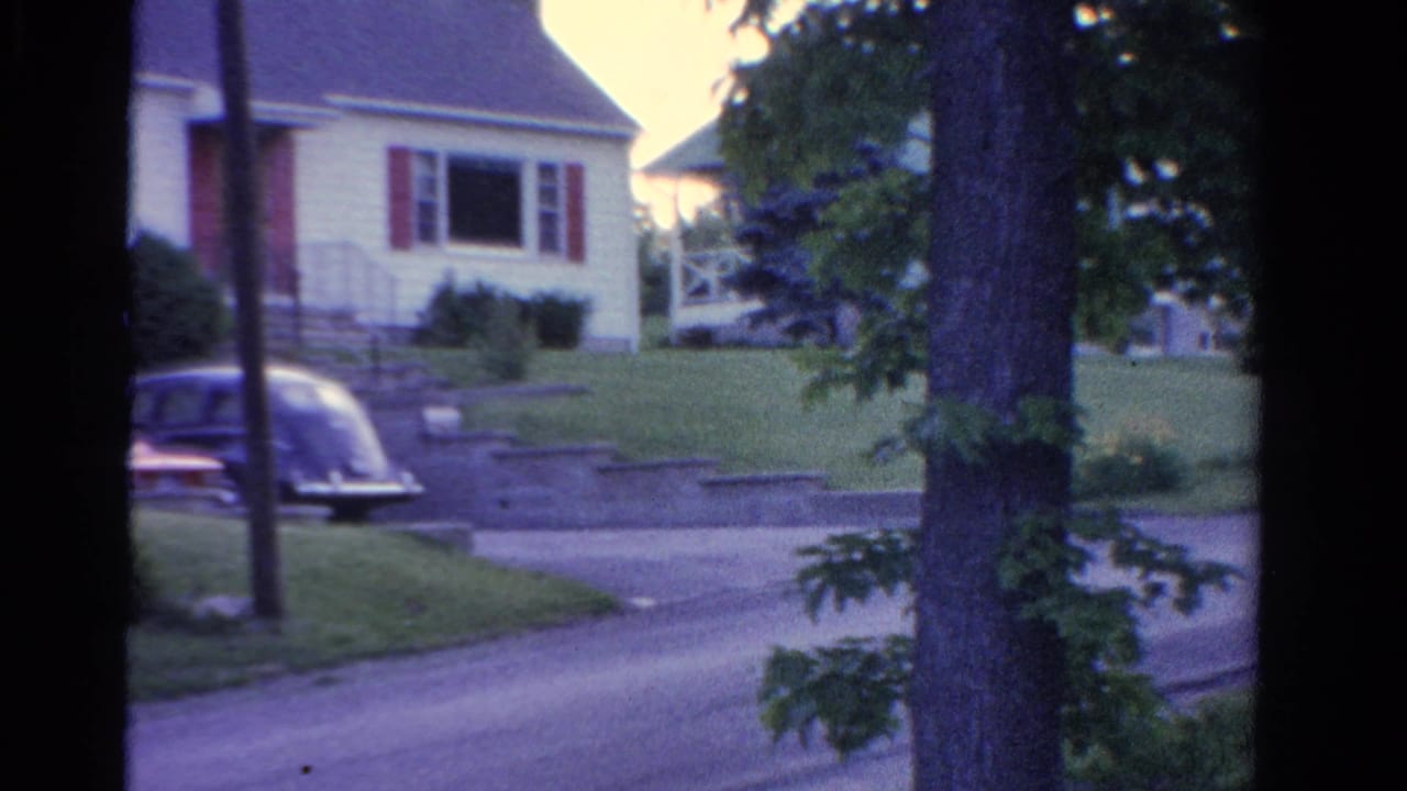 1958 Ontario residential view with a clapboard house, driveway, parked car, trimmed lawn, and trees seen through a dark window edge.