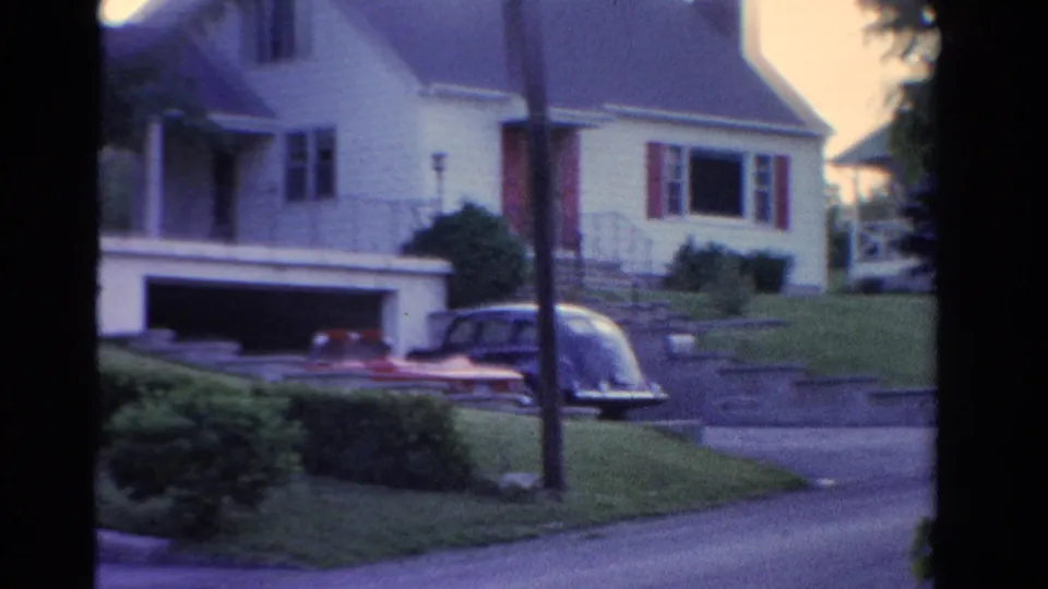 1958 Ontario home-movie frame showing a house with porch details, a driveway, a parked car, lawn, shrubs, and a roadside pole.