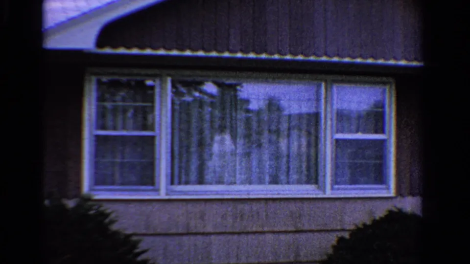 1958 Ontario 8mm close view of a residential window with curtains, exterior siding, roof awning, and dark shrubs in the foreground.