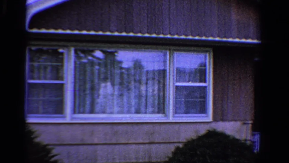 1958 Ontario 8mm frame showing a suburban house facade with brown siding, white windows, sheer curtains, and shrubs.