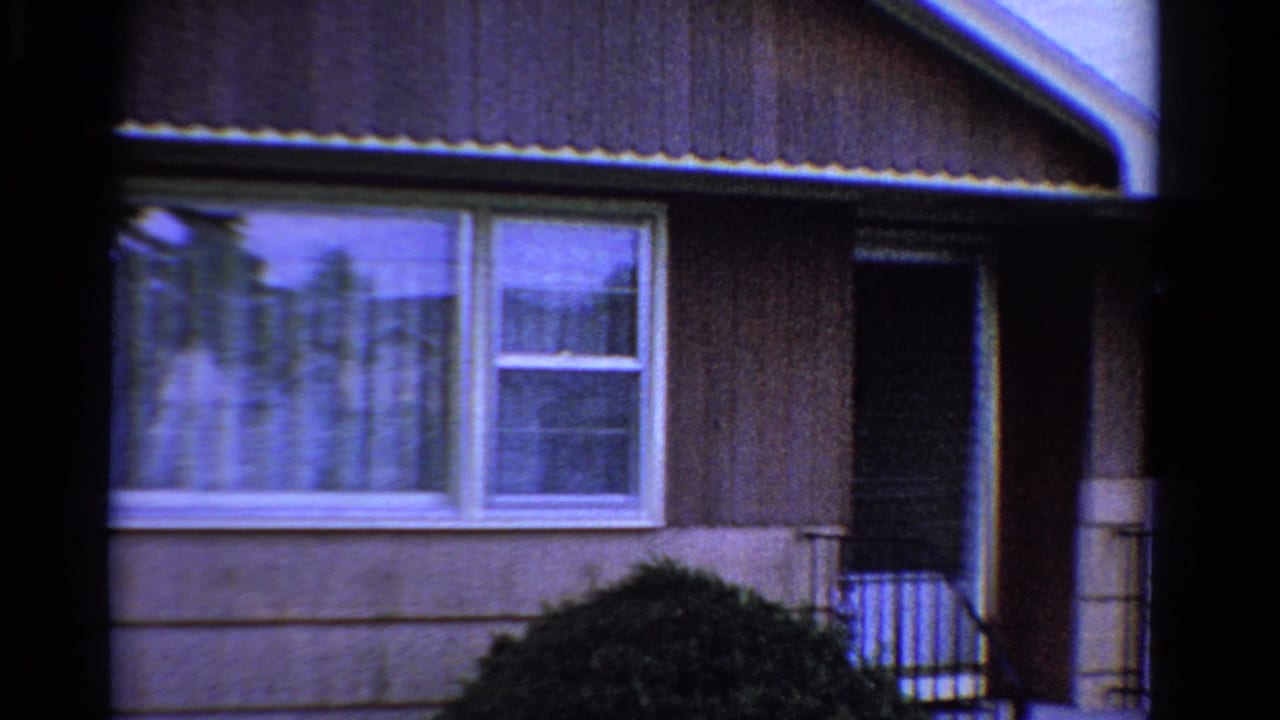 1958 Ontario home-movie frame with white windows, dark doorway, black railing, shrubs, and cool blue film cast.