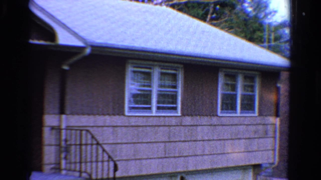 1958 Ontario home-movie frame showing a modest house facade with white windows, brown siding, pale roof, and a small porch railing.