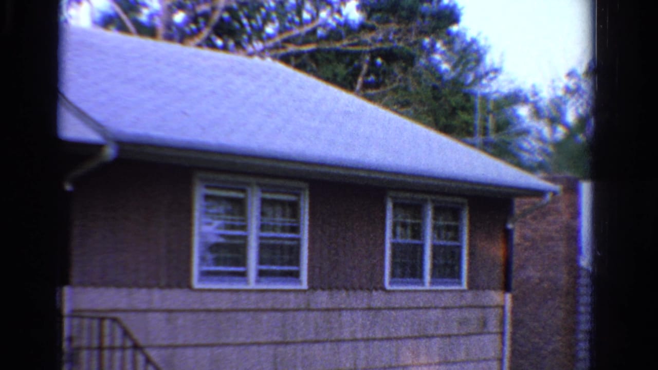 1958 Ontario home-movie view of the same modest house exterior, with white windows, brown siding, porch rail, roofline, and trees behind.