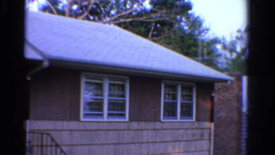 1958 Ontario home-movie view of the same modest house exterior, with white windows, brown siding, porch rail, roofline, and trees behind.