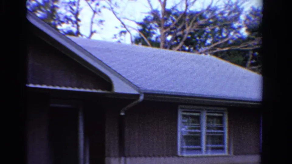 1958 Ontario frame with a closer low-angle view of the roofline, gable, brown siding, white windows, and trees behind the house.