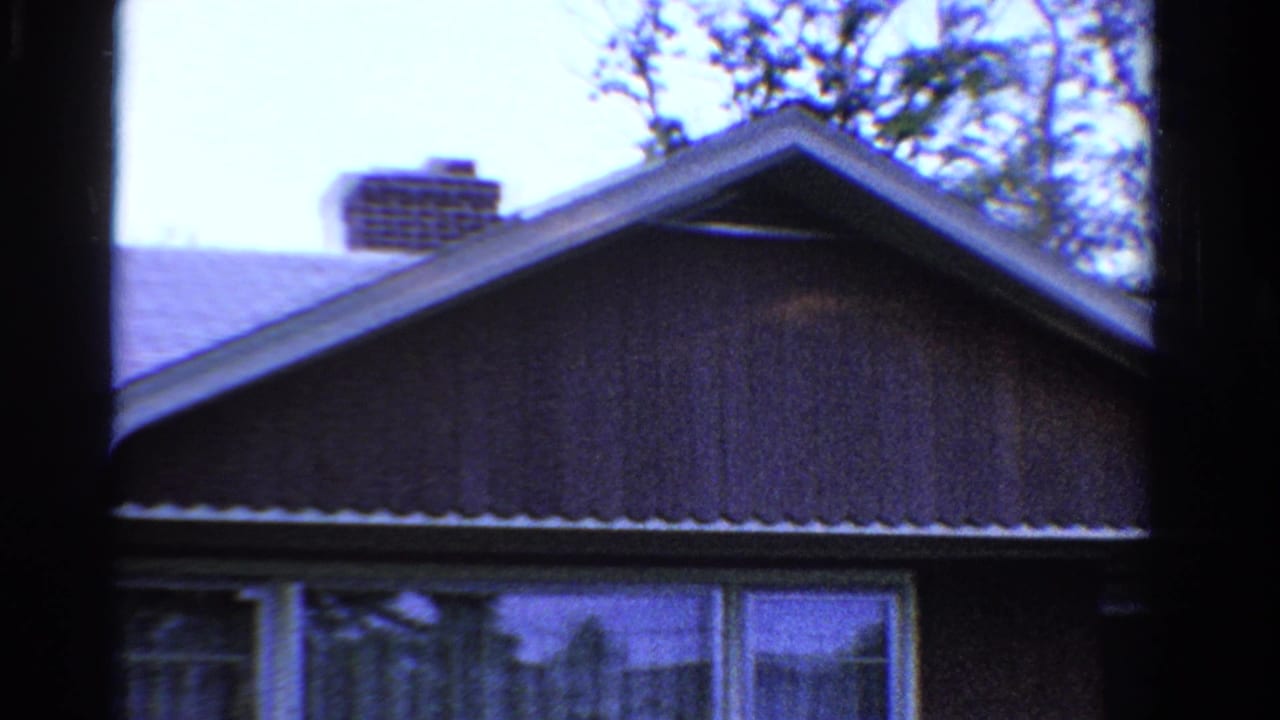 1958 Ontario home-movie frame showing a suburban house exterior with shingle roof, brick chimney, upper windows, and garage area.
