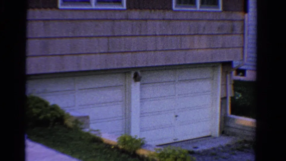 1958 Ontario home exterior with garage door, horizontal lower siding, upper windows, shrubs, and driveway foreground.
