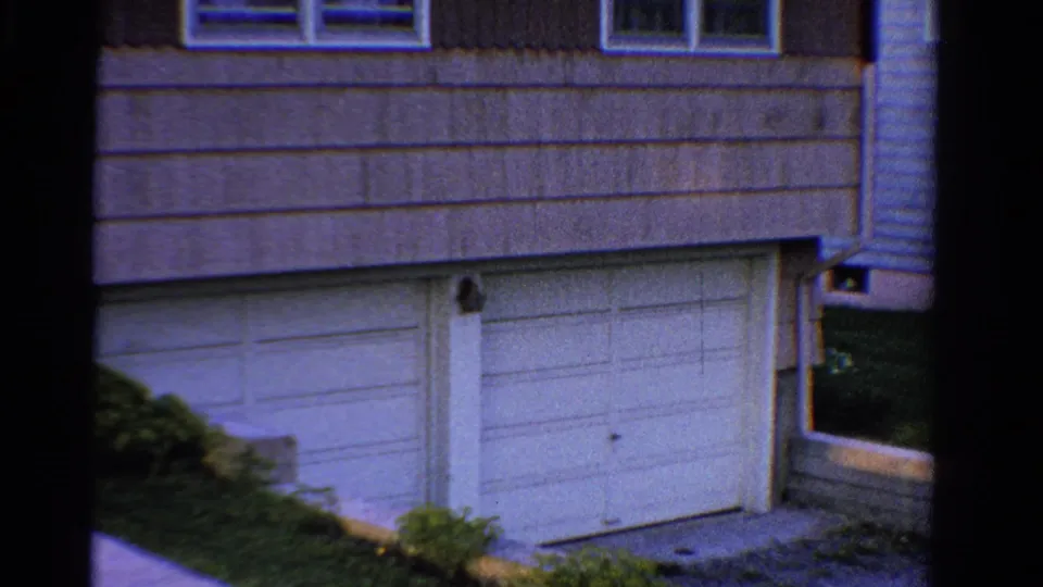 1958 Ontario frame showing the two-car garage facade of a suburban residence, with upper windows, siding, shrubs, and roofline.