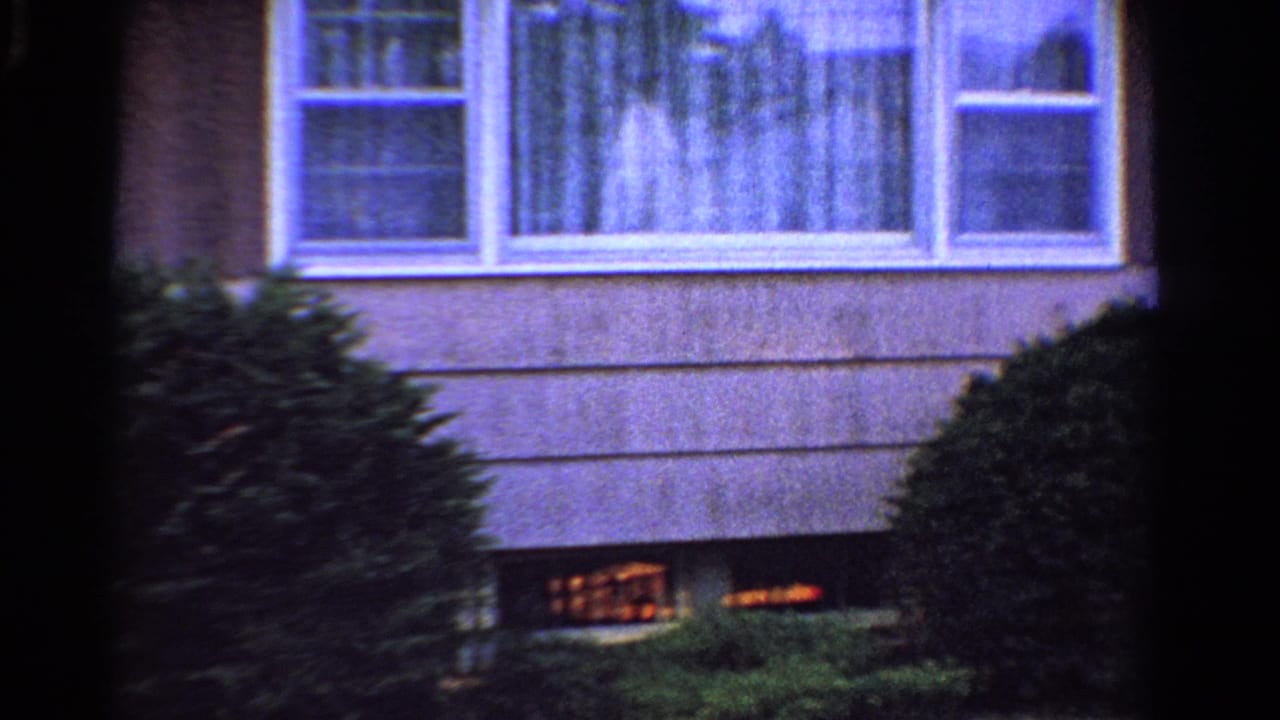 1958 Ontario residential exterior with curtained windows, shrubs, brown siding, lawn, and warm light near the foundation.