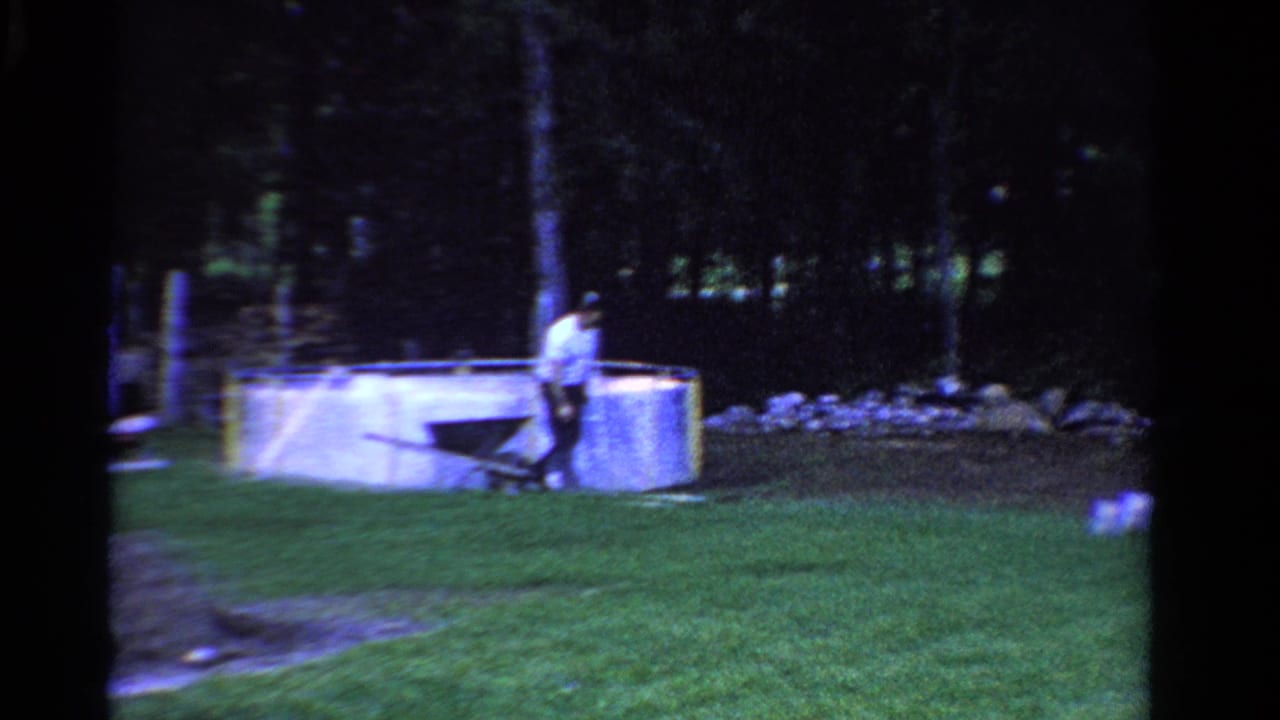 Man pushing a wheelbarrow near a small yard pool in a grassy wooded homestead.