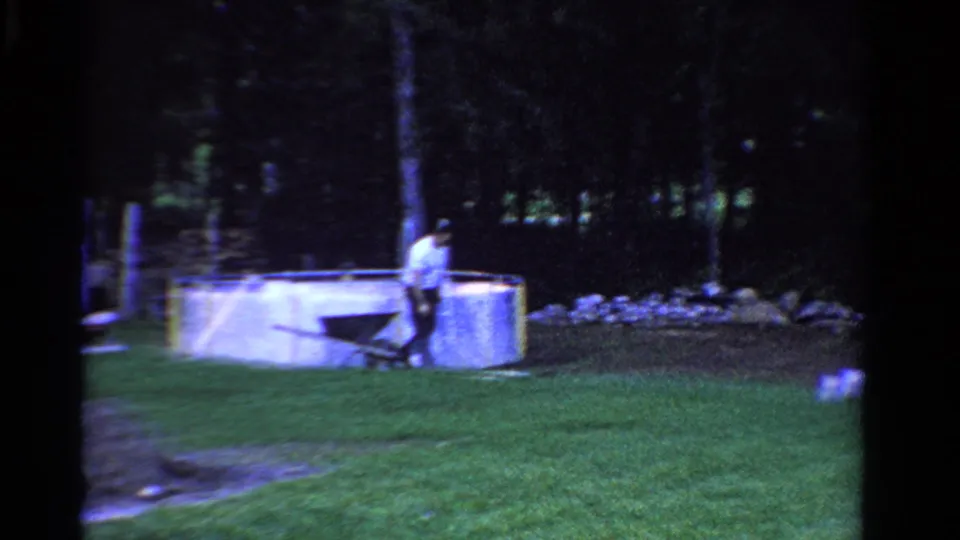 1958 Ontario home-movie frame showing a man with a wheelbarrow beside a small yard pool, with lawn and trees behind him.