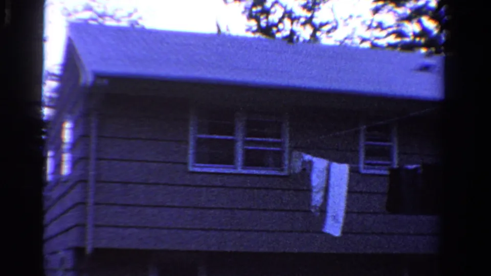 1958 Ontario home-movie frame showing a gray-sided house, pale roof, front windows, and laundry on a clothesline.