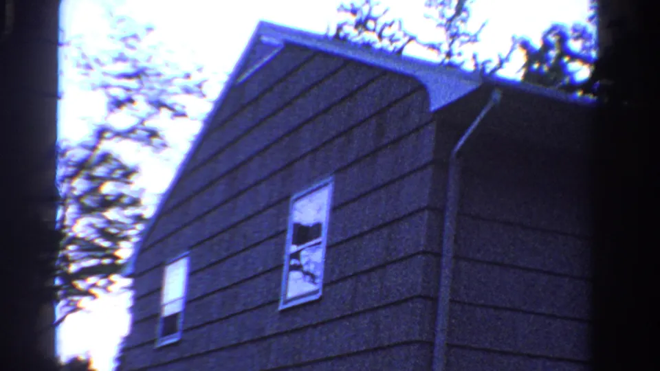 1958 Ontario home-movie frame showing the side wall of a gray house with two windows and trees beyond the roofline.
