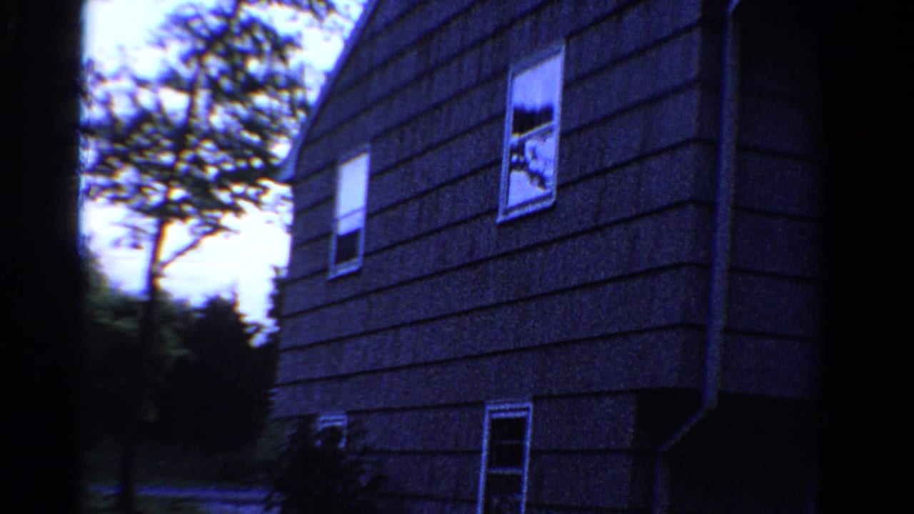 1958 Ontario home-movie frame of a shingled house side, with white-trimmed windows, trees, shrubs, and a quiet road nearby.