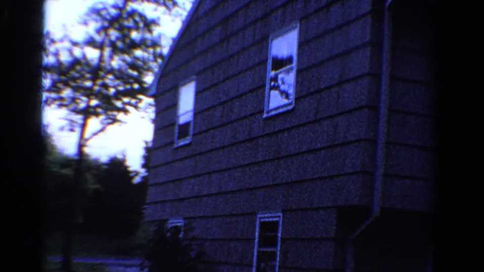 1958 Ontario home-movie frame of a shingled house side, with white-trimmed windows, trees, shrubs, and a quiet road nearby.