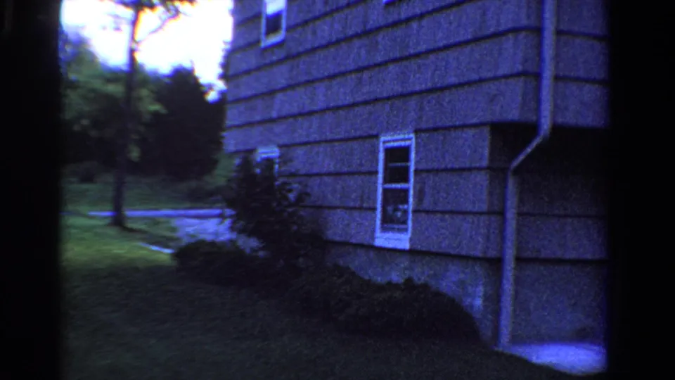 1958 Ontario 8mm view of the lower side of a shingled house, showing a downspout, basement window, shrubs, lawn, and roadside trees.