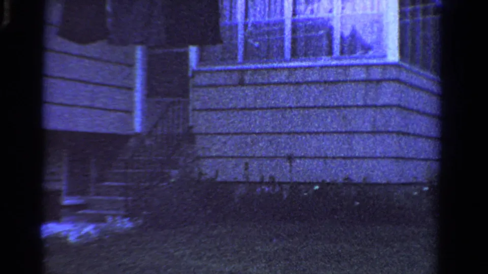 1958 Ontario frame showing the porch side of a shingled house, with railings, steps, white trim, shrubs, and blue-tinted 8mm grain.