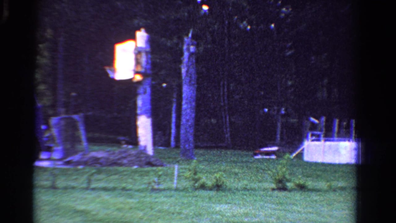 1958 Ontario 8mm frame showing a dusky backyard with grass, trees, a glowing yard light, and a small above-ground pool.