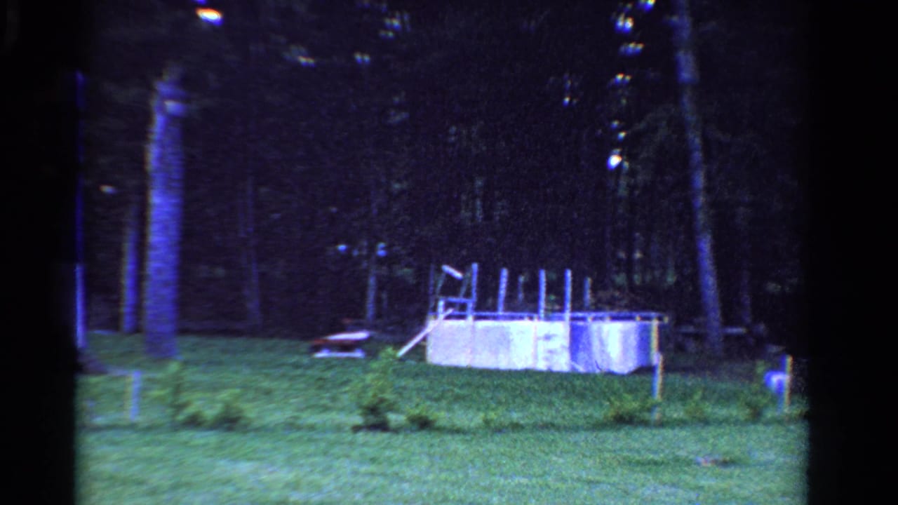 1958 Ontario home-movie frame of a suburban backyard with lawn, trees, a small white pool, and scattered outdoor objects.