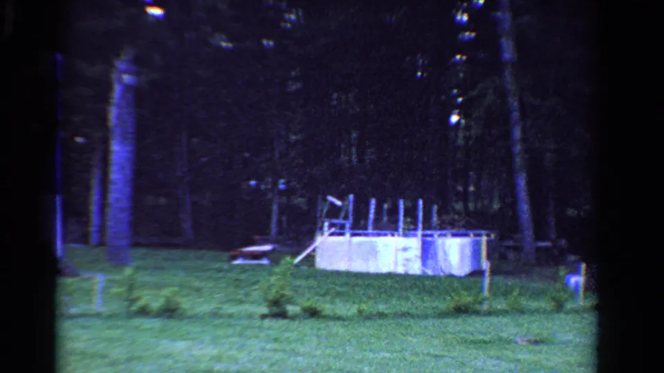 1958 Ontario home-movie frame of a suburban backyard with lawn, trees, a small white pool, and scattered outdoor objects.