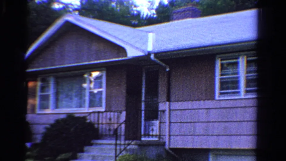 1958 Ontario home-movie frame of a house front with porch steps, railings, siding, windows, roofline, and dark doorway.