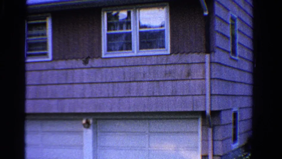 1958 Ontario home-movie frame showing a two-story suburban house facade with horizontal siding, upper windows, and white garage doors.