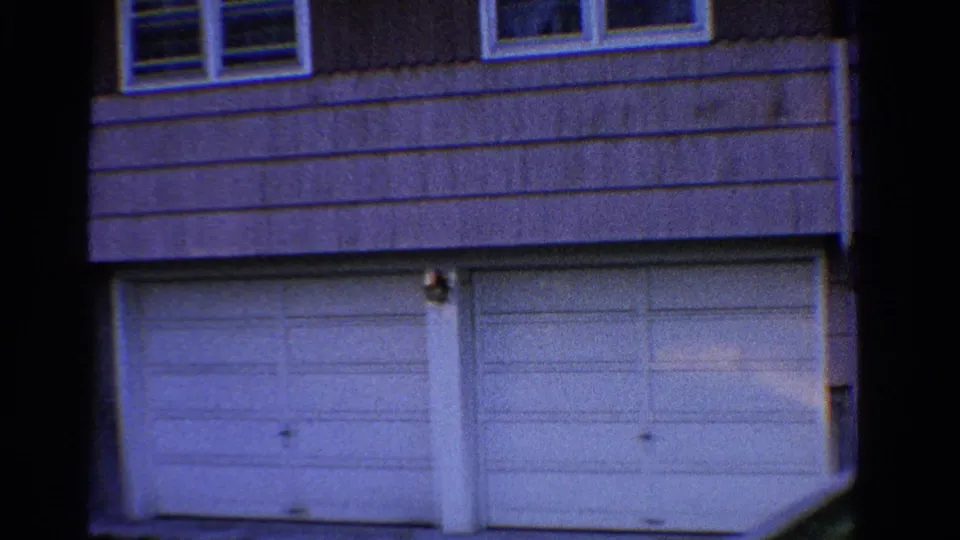 1958 Ontario home-movie frame with a closer view of white garage doors, horizontal siding, upper windows, and blue-tinted 8mm color.
