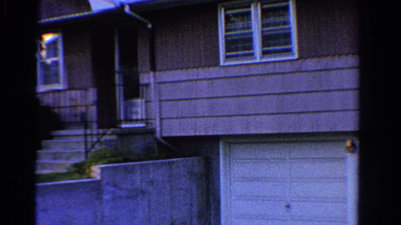 1958 Ontario home-movie frame showing a gray suburban house exterior with front steps, windows, garage, lawn, and low wall.