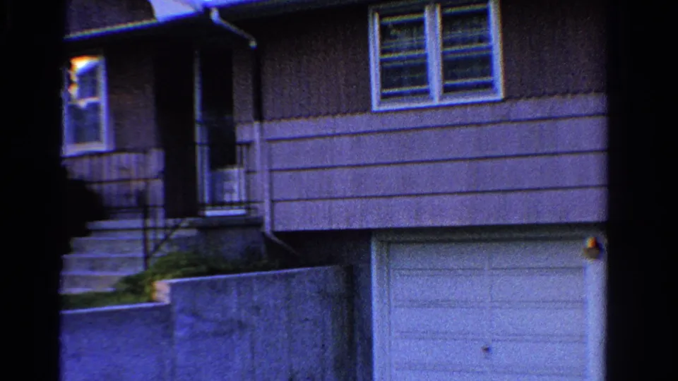 1958 Ontario home-movie frame showing a gray suburban house exterior with front steps, windows, garage, lawn, and low wall.