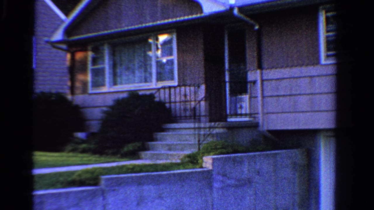 1958 Ontario frame of a gray suburban house entrance with steps, railings, front window, shrubs, lawn, and retaining wall.