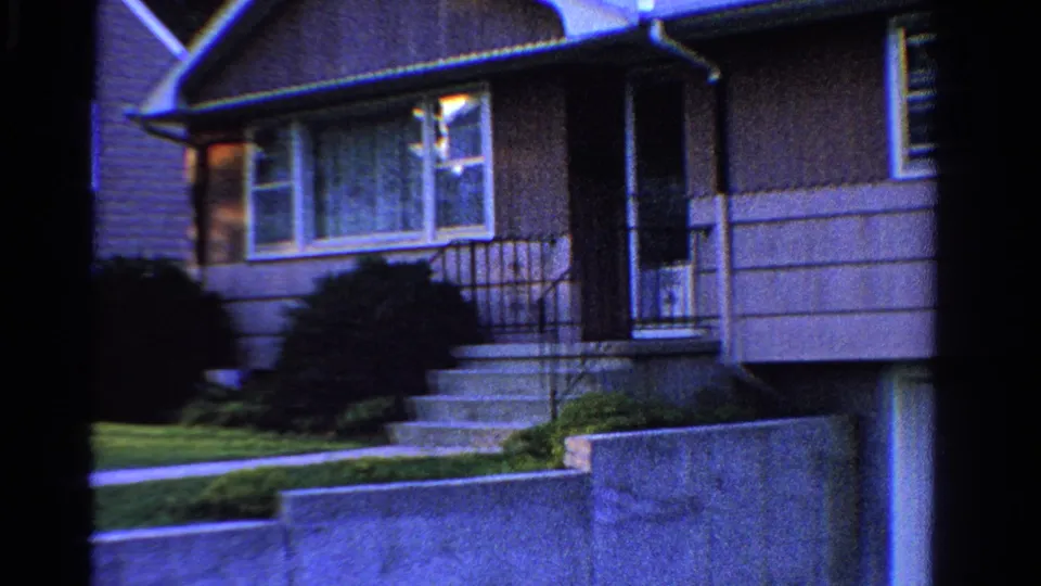 1958 Ontario frame of a gray suburban house entrance with steps, railings, front window, shrubs, lawn, and retaining wall.