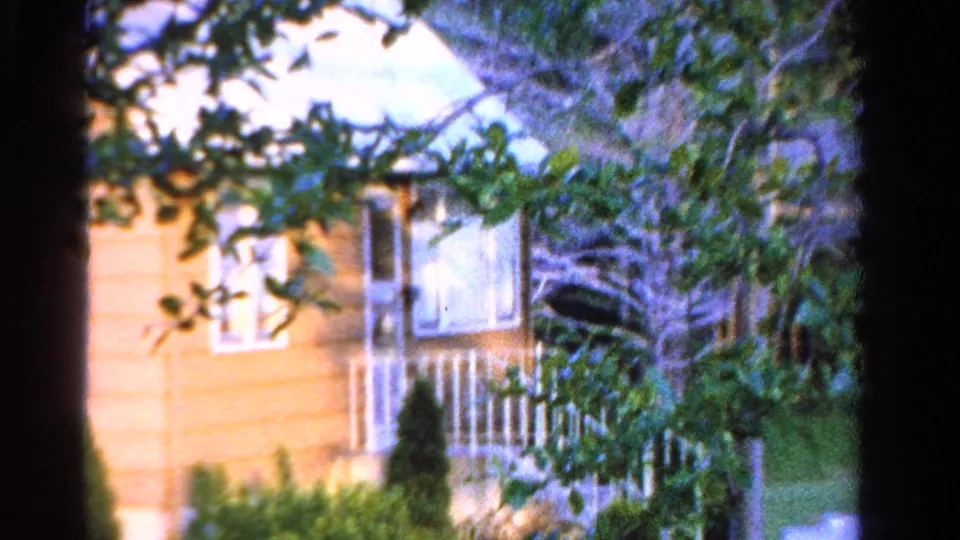 1958 Ontario home-movie frame showing a brown suburban house partly obscured by tree branches, with windows, steps, railing, shrubs, and lawn.