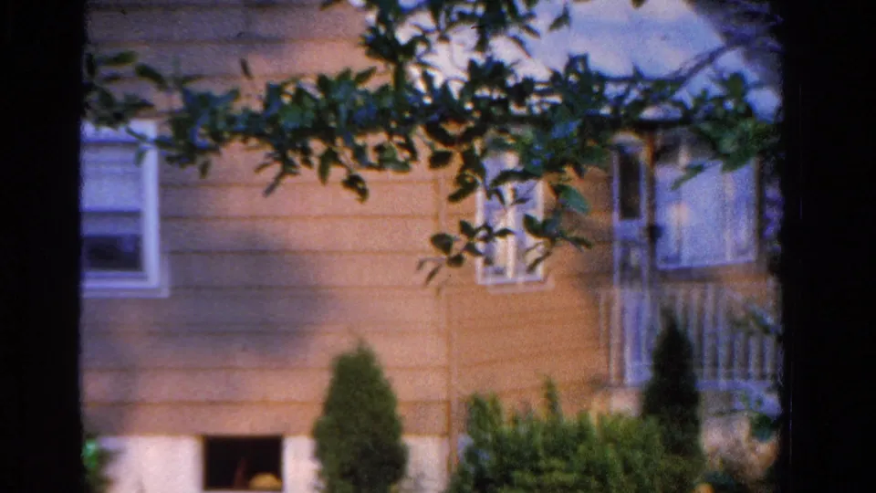 1958 Ontario home-movie frame of a light-colored suburban house seen through tree trunks and leaves.