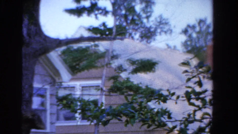 1958 Ontario frame showing a suburban roofline and upper windows through trees in soft 8mm color.