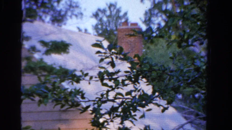 1958 Ontario home-movie frame with a sloped roof, brick chimney, and leafy branches in the foreground.