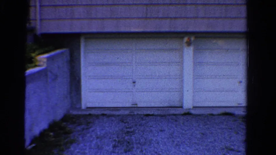 1958 Ontario frame with white paneled garage doors, gravel ground, and a low retaining wall.