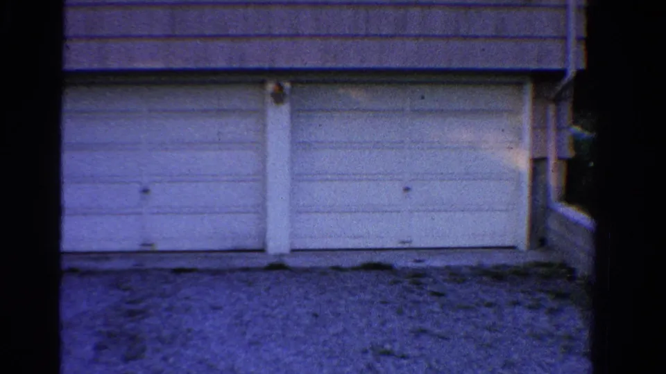 1958 Ontario home-movie frame showing the garage facade with paneled doors and gravel in front.