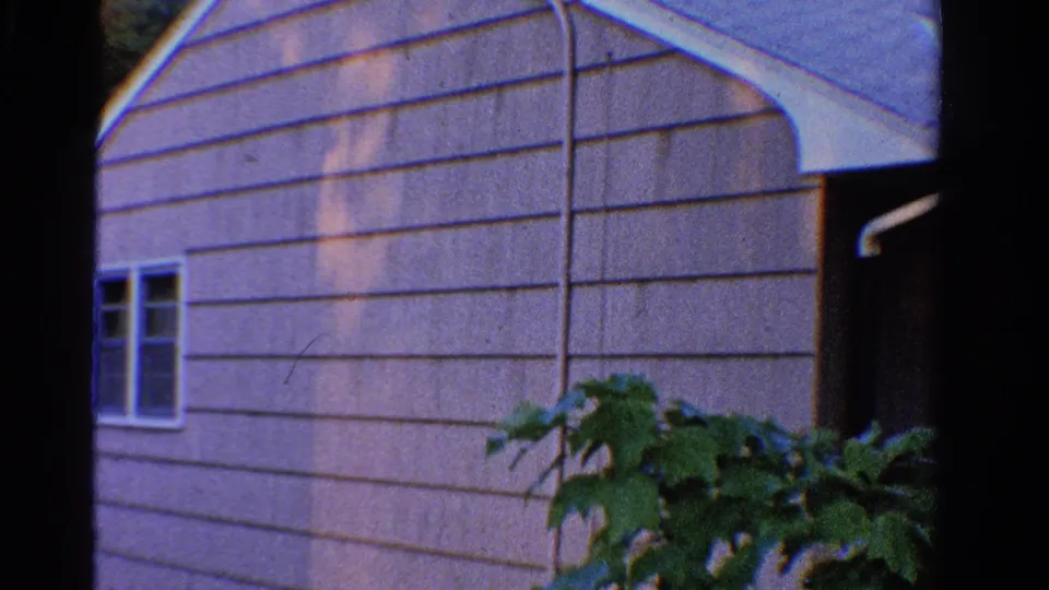1958 Ontario home-movie frame showing the side of a light-sided house with windows, plants, and adjacent rooflines.
