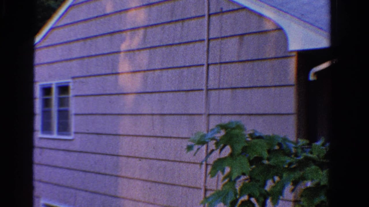 1958 Ontario 8mm frame showing a light-sided house exterior, white-trimmed windows, and leafy foreground plant.