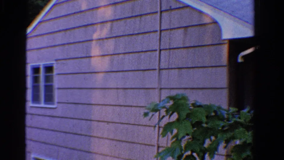 1958 Ontario 8mm frame showing a light-sided house exterior, white-trimmed windows, and leafy foreground plant.
